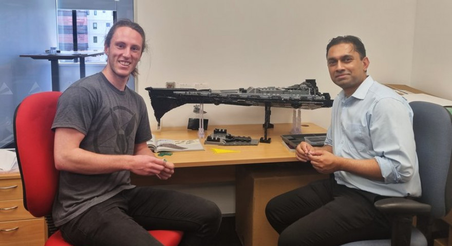 Liam Sharpe and Sanjay DSouza smiling are sitting on chairs in a brightly lit office with a large unfinished Star Wars Lego set on the table behind them.